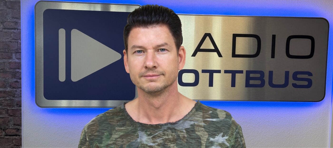 DJ Enrico Ostendorf steht vor dem Logo von Radio Cottbus