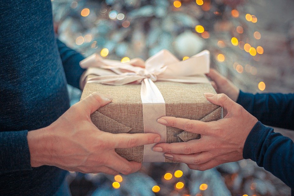 Geschenk wird übergeben, im Hintergrund ein leuchtender Tannenbaum, das Geschenk ist beige mit Schleife