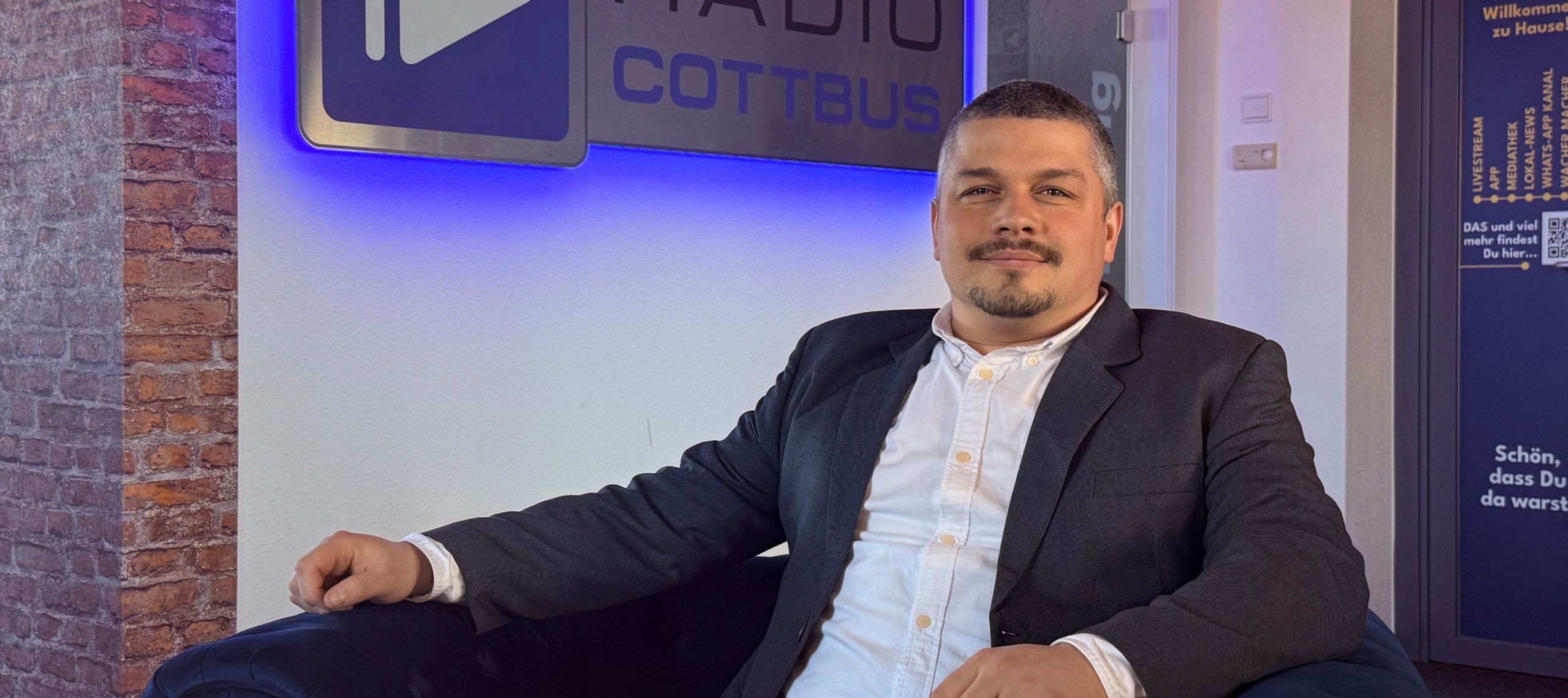 Tim Fielo Mediaberater von Radio Cottbus sitzt vor dem Logo von Radio Cottbus