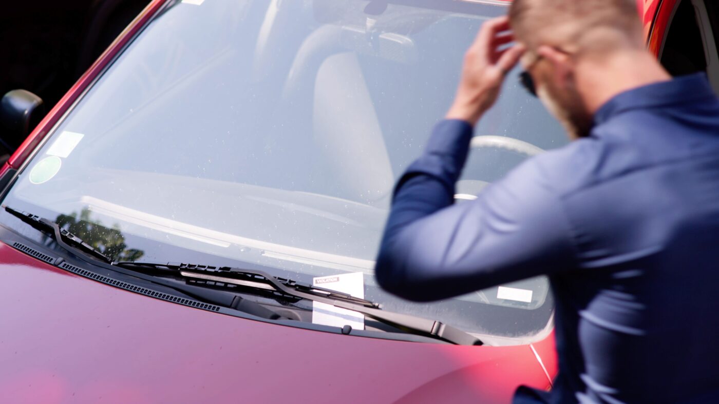 Mann steht vor seinem Auto und schaut auf einen Strafzettel, der an seinem Scheibenwischer. Er ärgert sich und schlägt die Hand vor den Kopf.