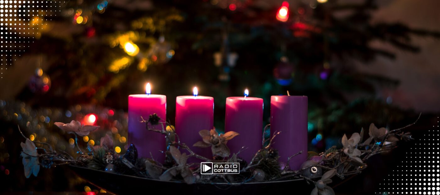 4 Kerzen in einem Adventsgesteck vor einem geschmückten Weihnachtsbaum