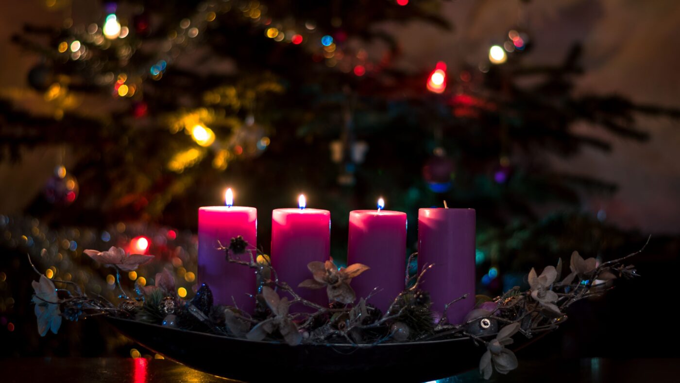 4 Kerzen in einem Adventsgesteck vor einem geschmückten Weihnachtsbaum