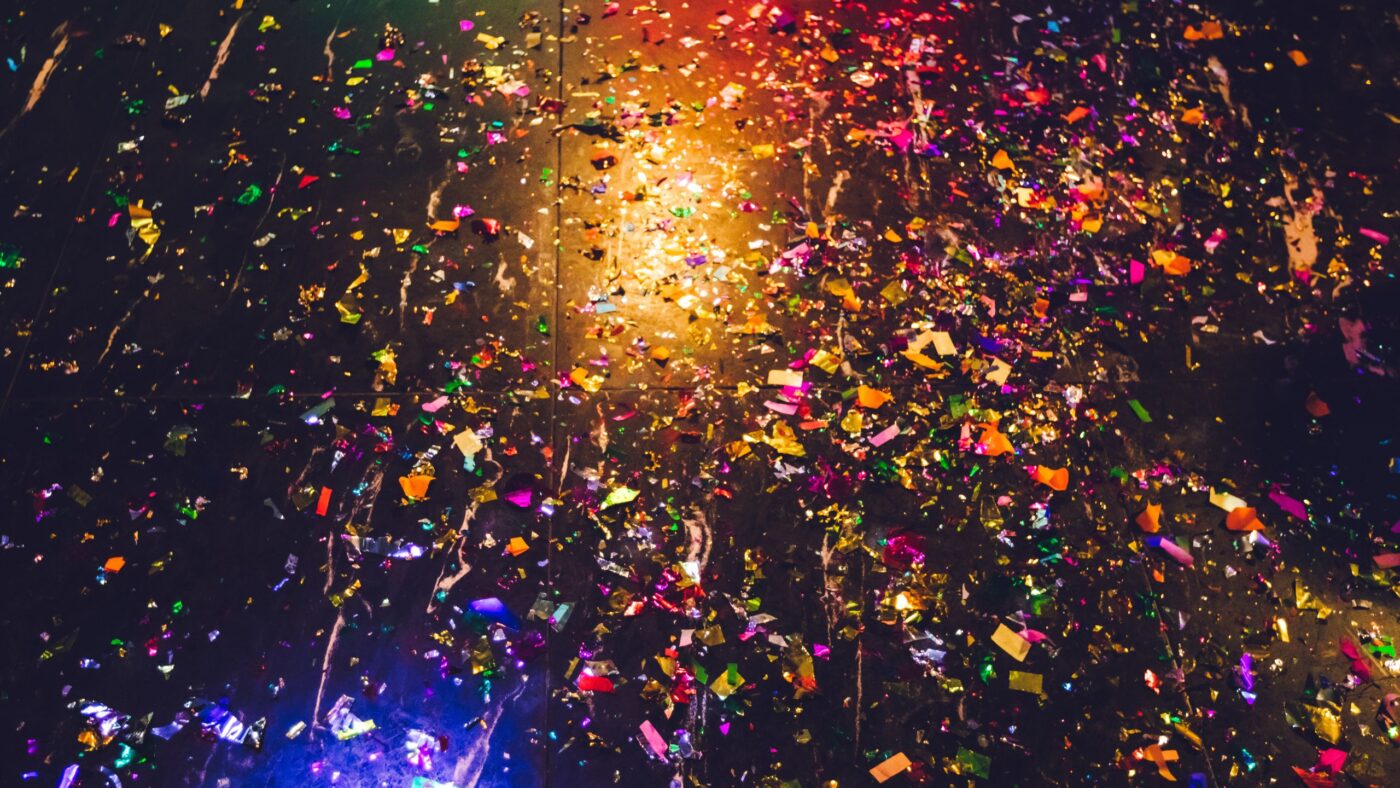 buntes Konfetti vor einem gelben und einem blauen Lichtscheinwerfer auf schwarzem Hintergrund.