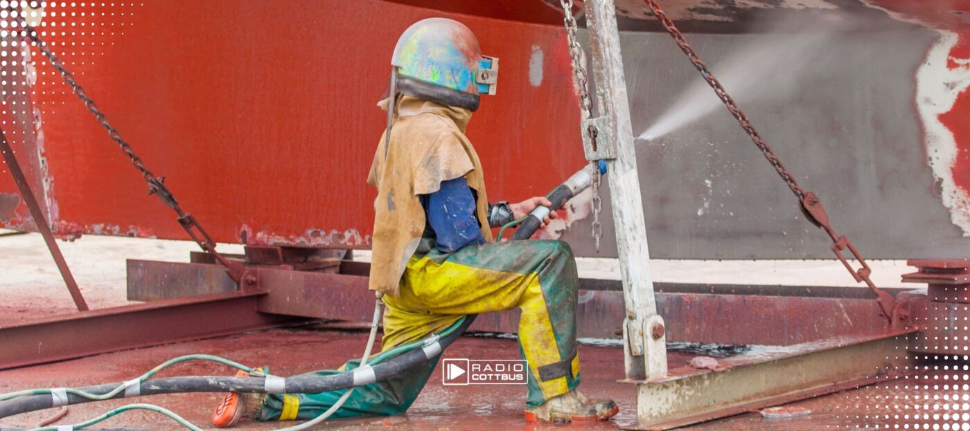Ein Mann in Schutzkleidung wäscht mit einem Wasserschlauch Farbe von einem Gebäude ab.