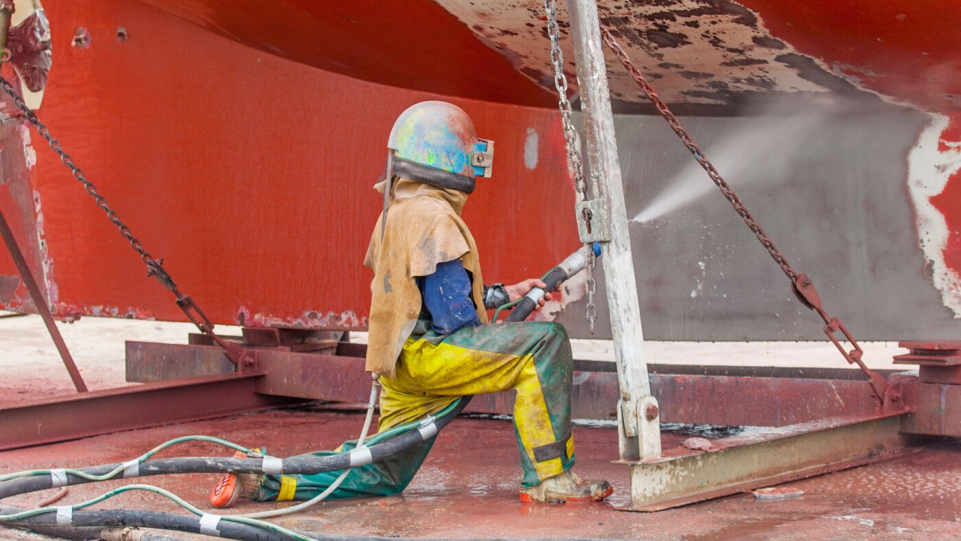 Ein Mann in Schutzkleidung wäscht mit einem Wasserschlauch Farbe von einem Gebäude ab.