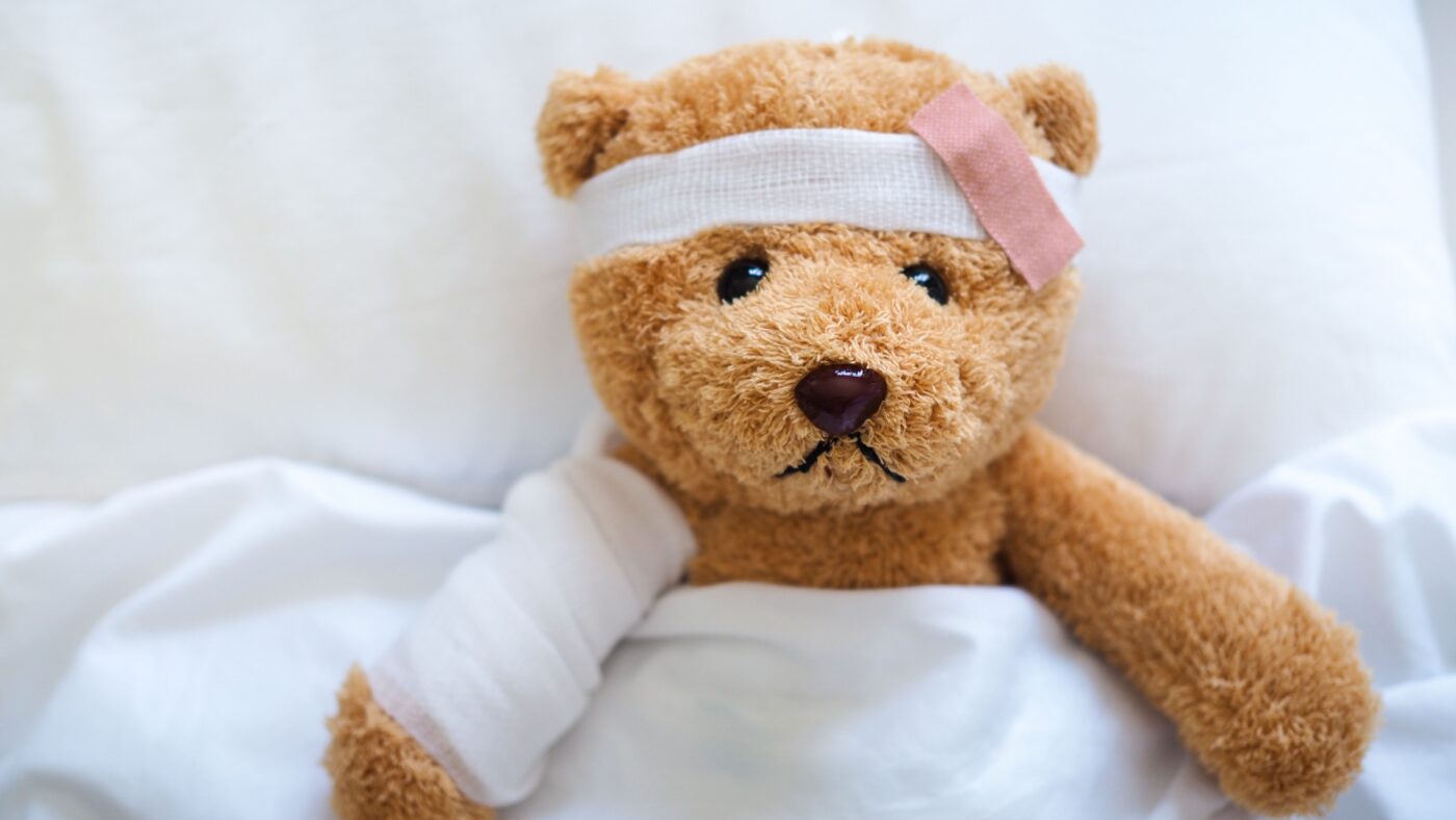 Ein brauner Teddybär liegt zudeckt in einem Bett, er hat Verbände um den rechten Arm und um den Kopf. Auch ein Pflaster klebt auf seinem Kopf.