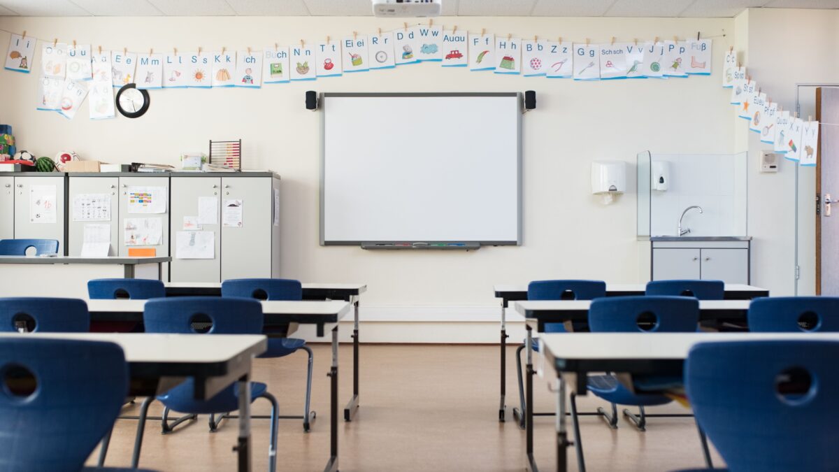 Ein leeres Klassenzimmer. Darin stehen Schulbänke mit Stühlen, vorne ist eine weiße Tafel