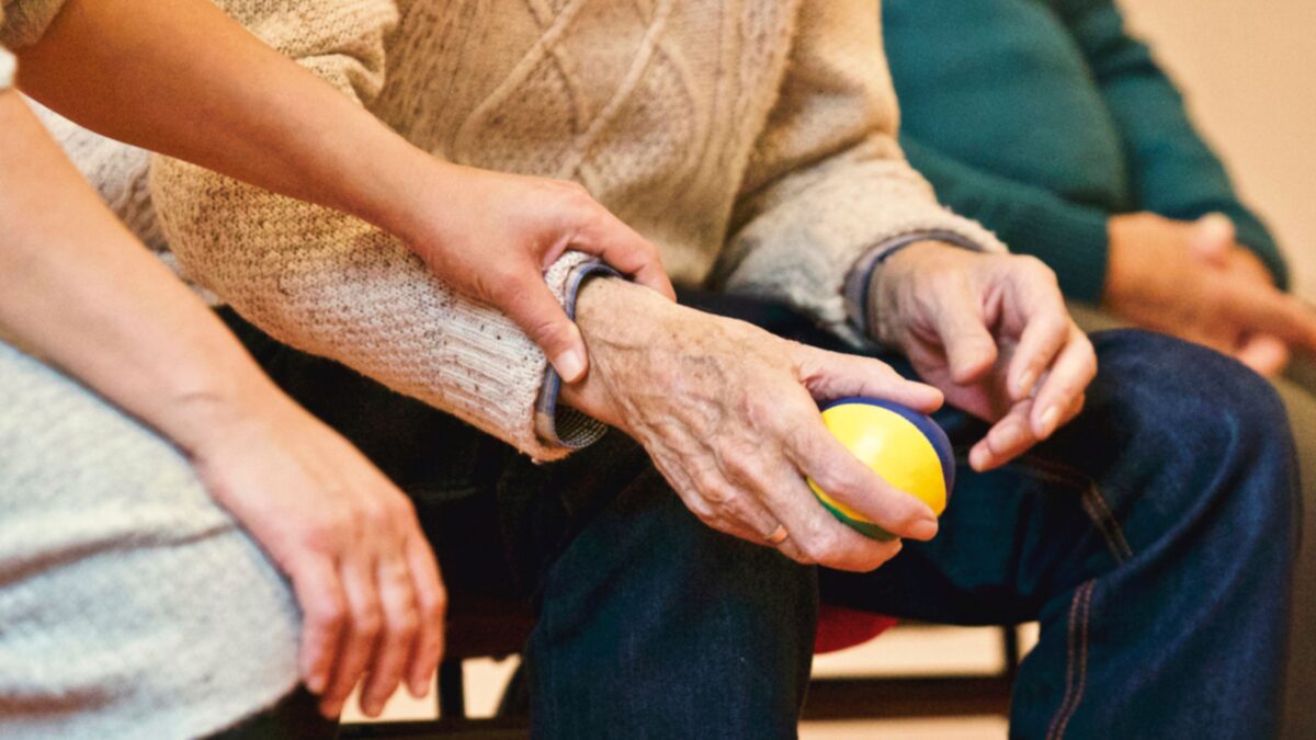 Man sieht drei Menschen nebeneinander, Schulterabwärts, in der Mitte sitzt ein älterer Mann, der einen Ball in den Händen hält und daneben ist eine jüngere Frau, die ihm hilft