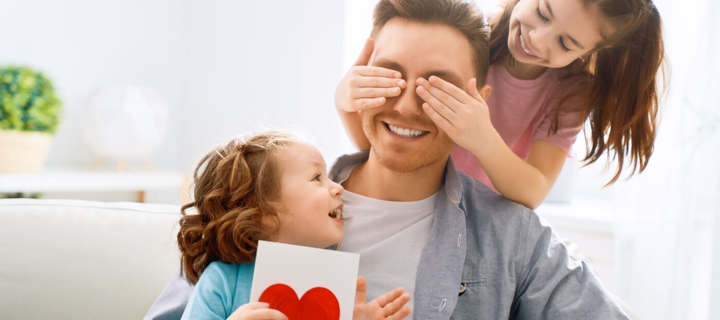 Zwei Kinder gratulieren ihrem Vater und eines hält ihm dabei die Augen zu, während das andere kleinere Kind ihm eine selbstgebastelte Glückwunschkarte mit einem Herz darauf schenk