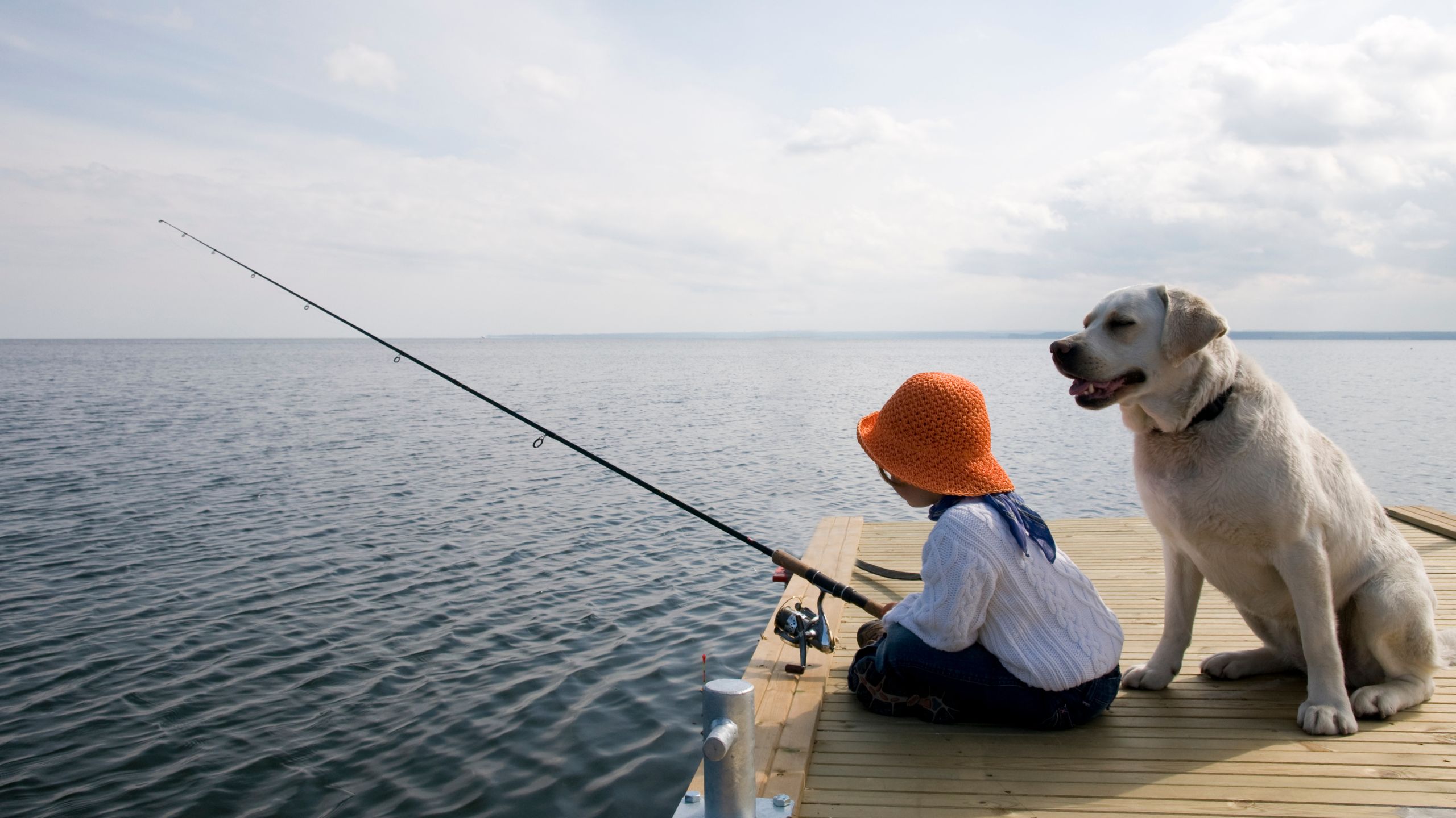Ein Kind und ein Hund sitzen auf auf einem Steg, das Kind hat eine Angel in der Hand. Im Hintergrund sieht man einen See mit weitem Horizont und darüber Wolken.