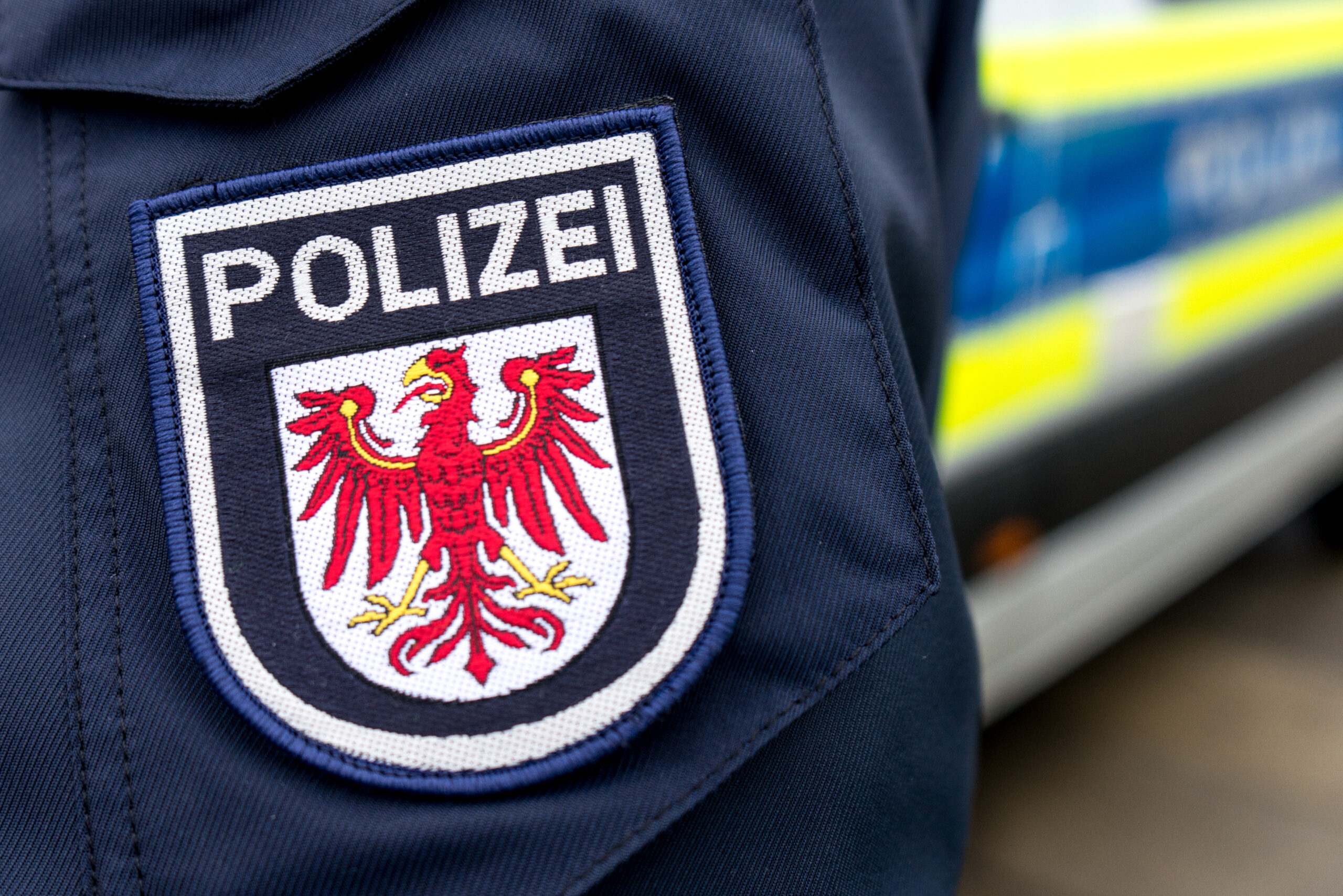 Ein Polizist vor einem Polizeiauto bei dem der Fokus auf das logo der Polizeijacke liegt