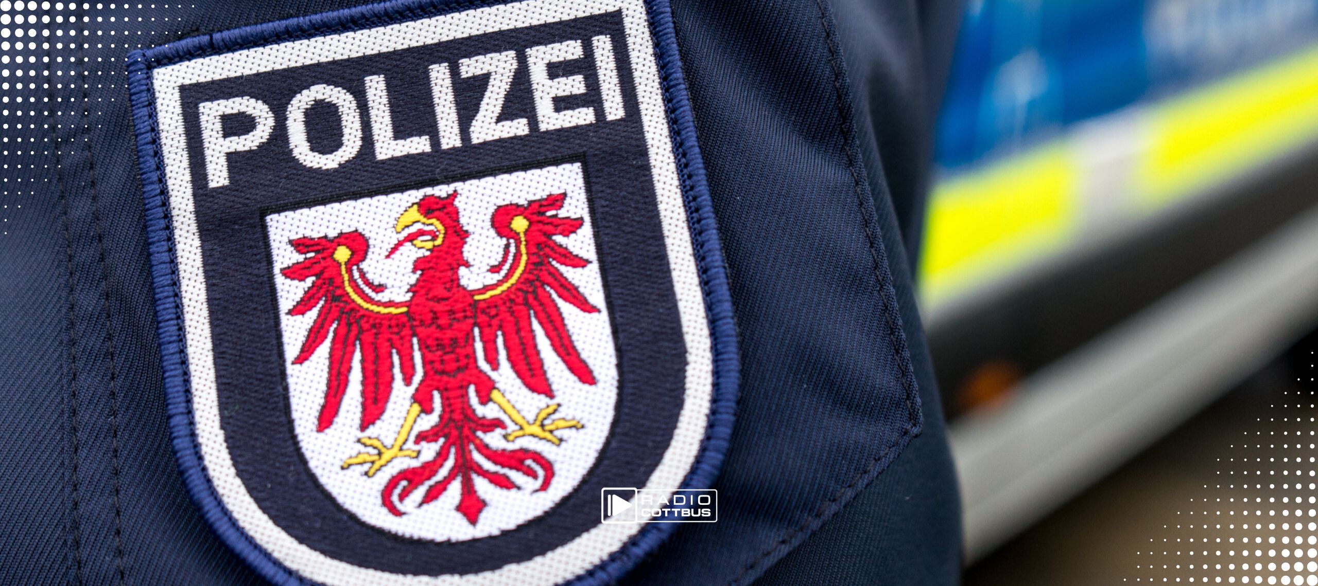 Ein Polizist vor einem Polizeiauto bei dem der Fokus auf das logo der Polizeijacke liegt