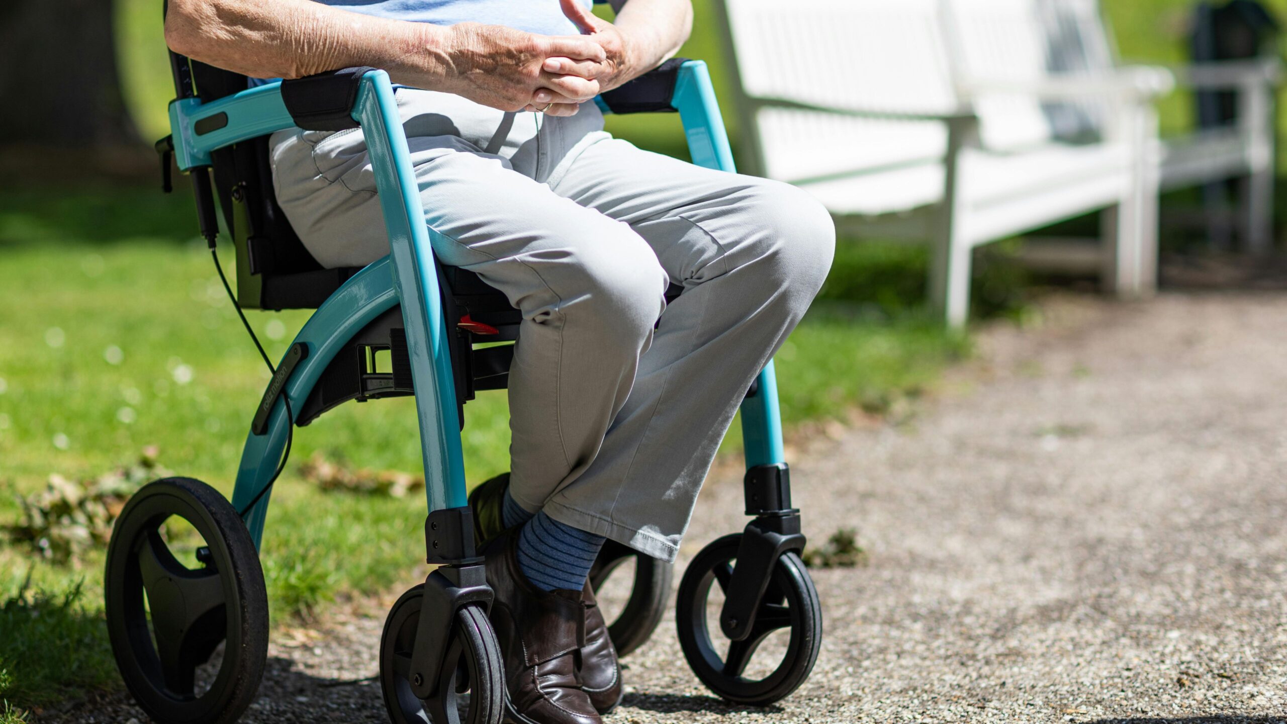 Ein älterer Mann sitzt auf einem Rollator neben einer weißen Parkbank