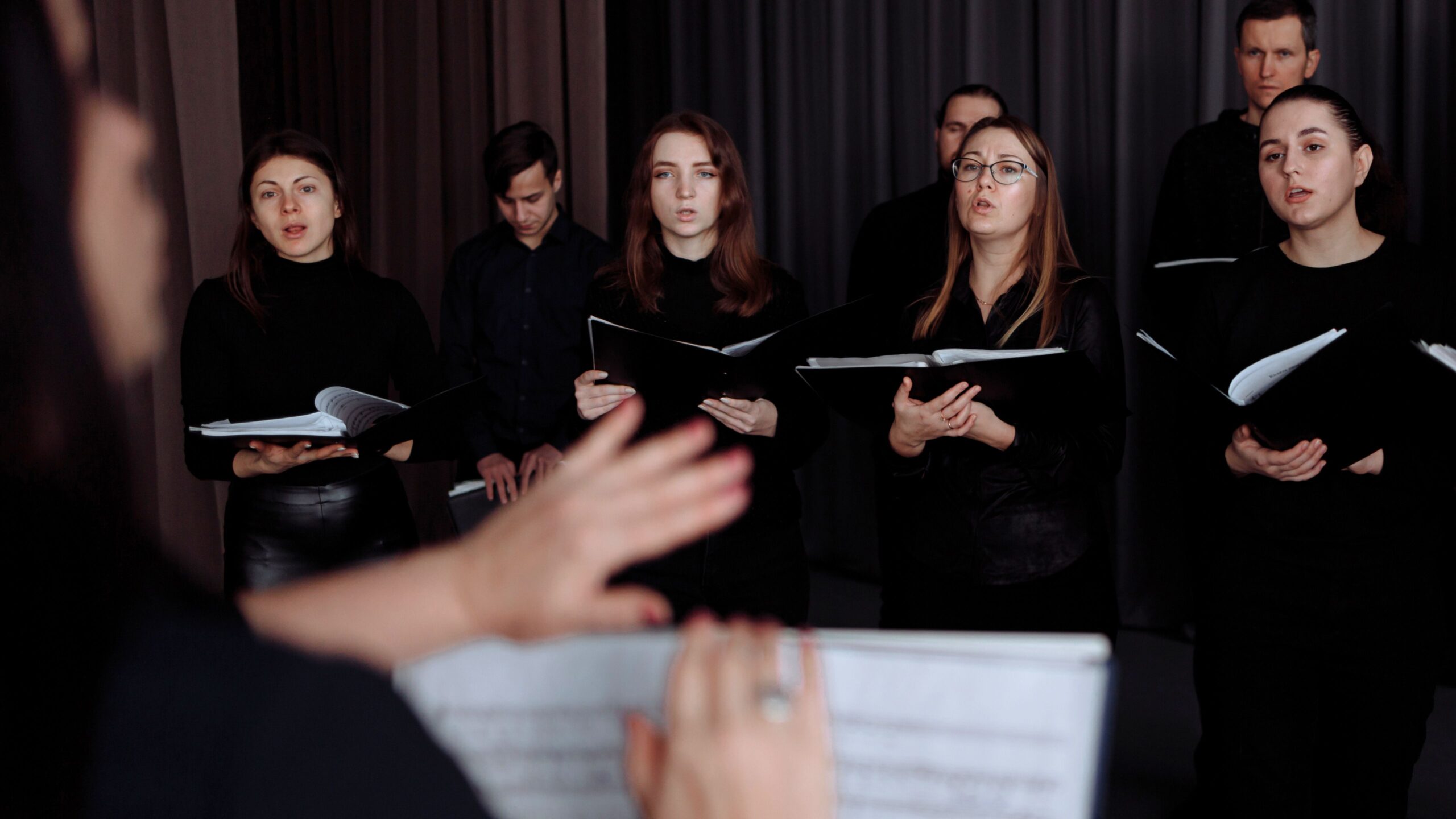 ein gemischter Chor mit vier Frauen in der ersten Reihe und drei Männern in der hinteren Reihe, unscharf im Vordergrund ist de Chorleiterin mit Notenblättern