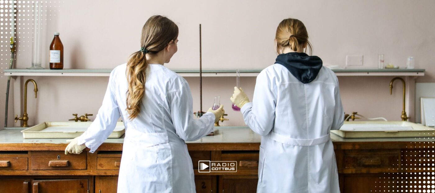 Zwei Frauen in Laborkitteln stehen an einem Tisch und experimentieren mit Gläsern