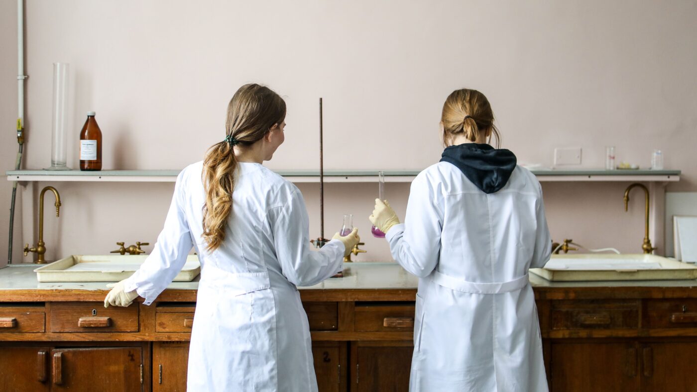 Zwei Frauen in Laborkitteln stehen an einem Tisch und experimentieren mit Gläsern