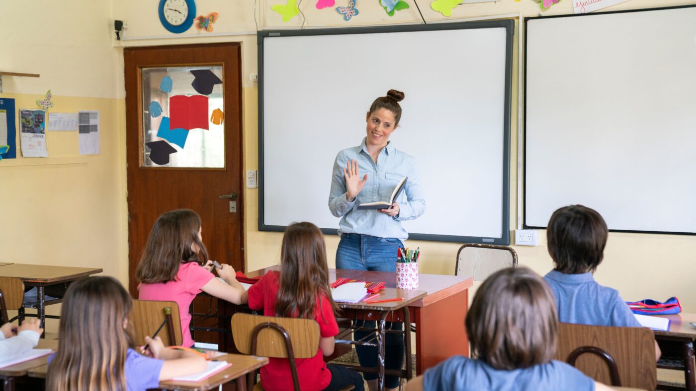Eine Lehrerin steht vor einer weißen Tafel und spricht zu ihrer Klasse