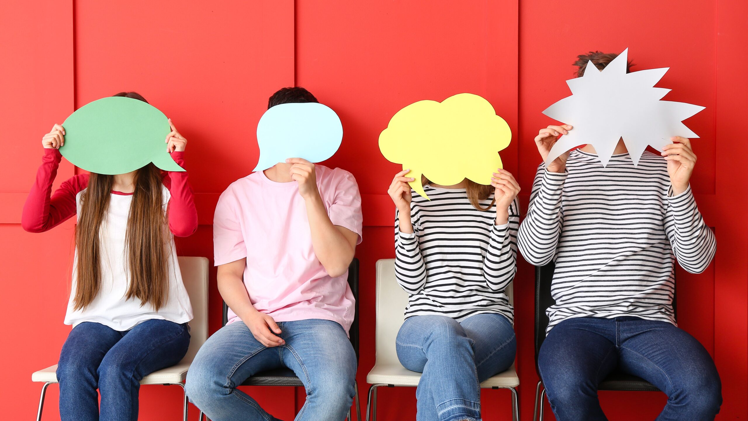 Vier Personen sitzen vor einem roten Hintergrund nebeneinander auf einer Bank und halten Schilder in unterschiedlichen Formen hoch. Die ersten beiden Personen von links halten eine Sprechblase in grün und hellblau. Rechts davon eine Denkblase in gelb und ganz rechts eine zackige Sprechblase.