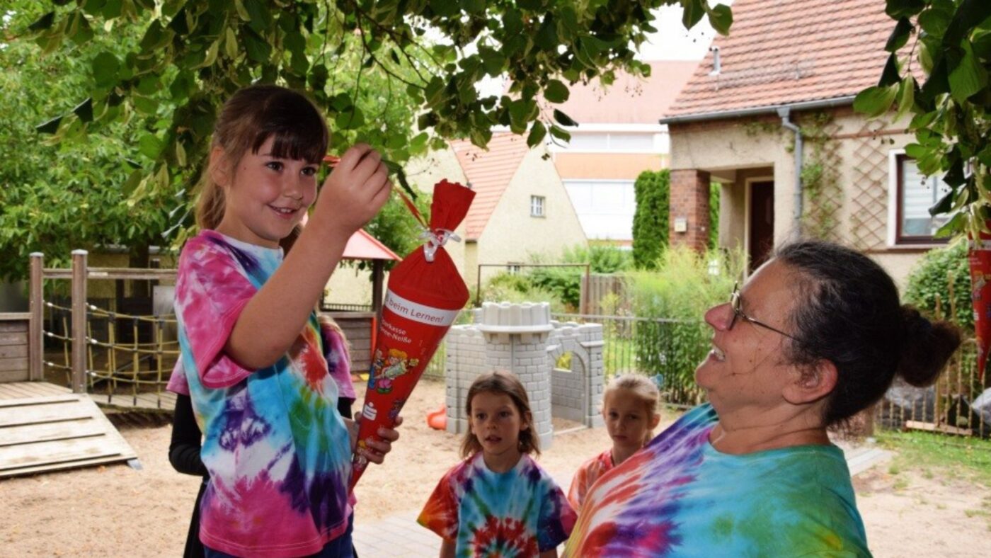 Mia Marie Kubens mit Erzieherin Ann-Marei Bischoff hängen gemeinsam eine Zuckertüte an einen Baum. Im Hintergrund sind andere Kinder, die dabei zuschauen.