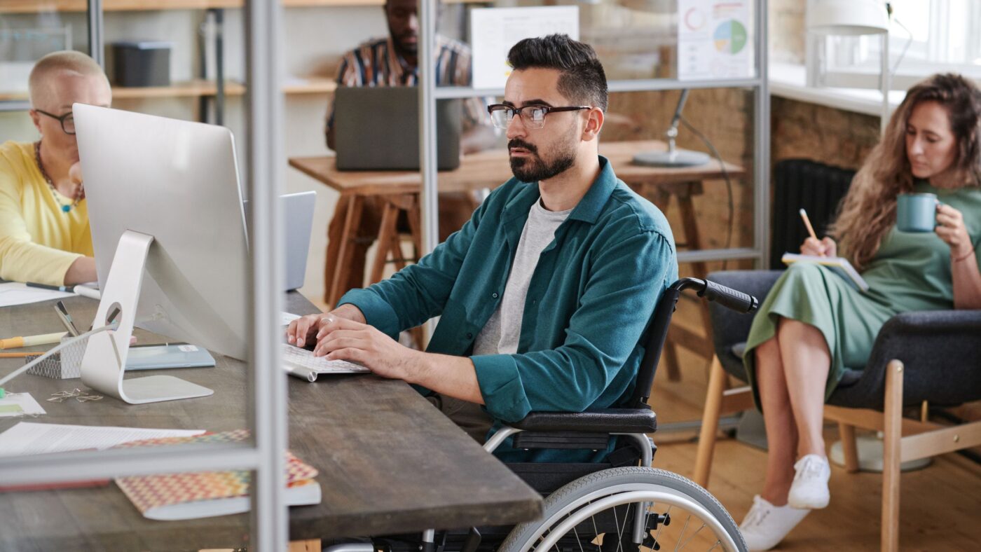 Man sieht ein Mann im Rollstuhl, welcher an seinem Arbeitsplatz sitzt und etwas am PC macht