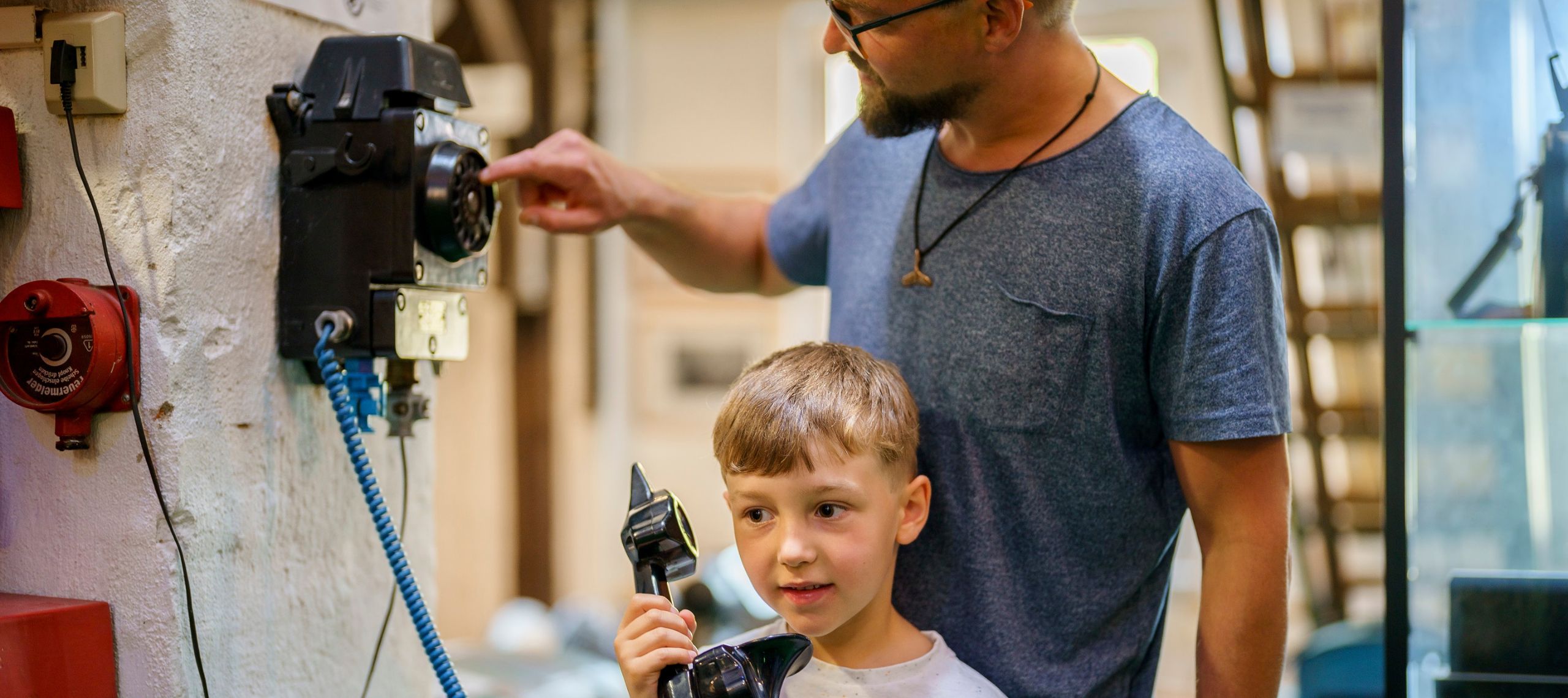 Ein kleiner Junge nimmt an einer Veranstaltung im Lübbener Ferienkalender teil und hat einen historischen Telefonhörer in der Hand während sein Vater hinter ihm steht und die Wählscheibe bedient