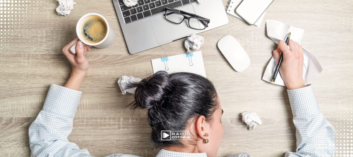 Person mit schwarzen Haaren und Dutt liegt auf dem Tisch, um sie rum Papier, ein Laptop, ein Kaffee und ein Stift. Die Aufnahme wurde von oben gemacht.