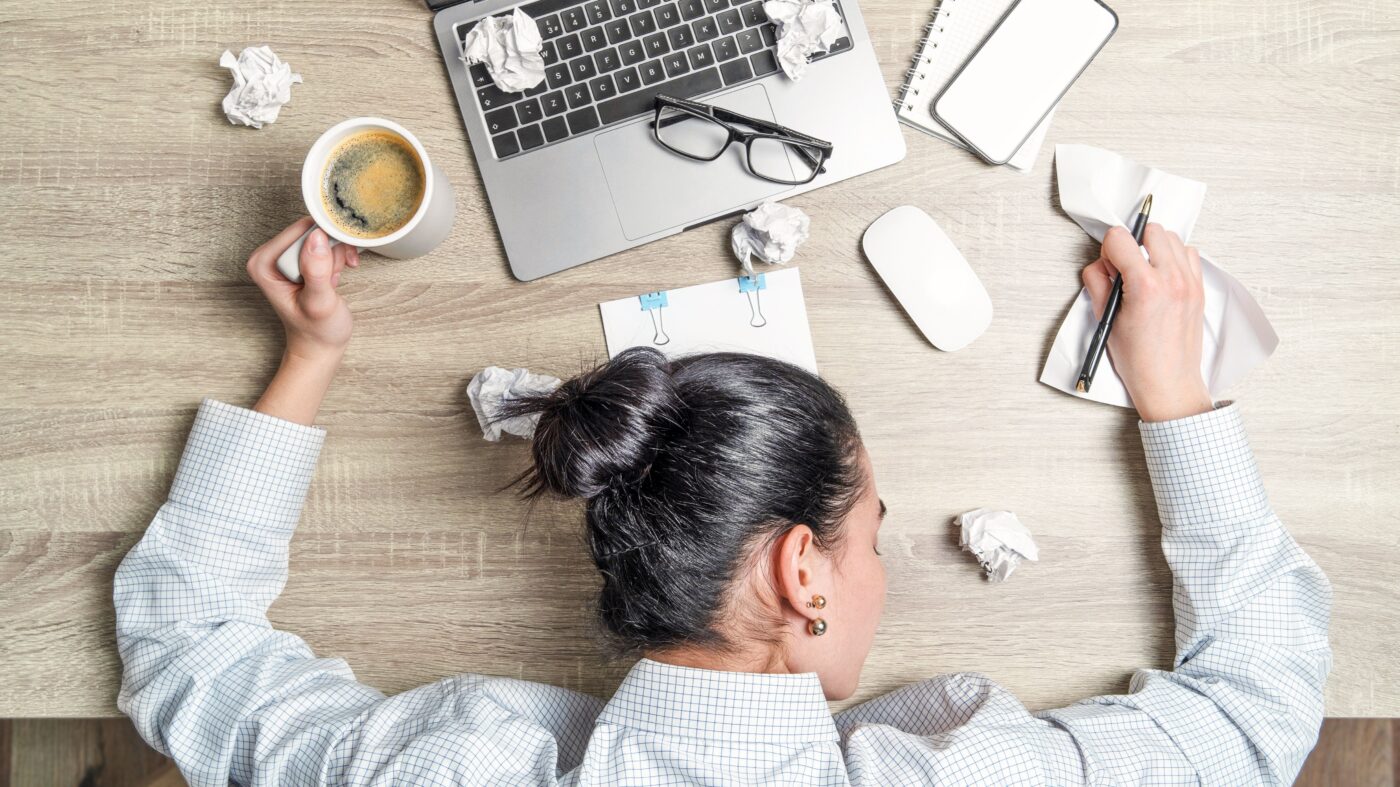Person mit schwarzen Haaren und Dutt liegt auf dem Tisch, um sie rum Papier, ein Laptop, ein Kaffee und ein Stift. Die Aufnahme wurde von oben gemacht.