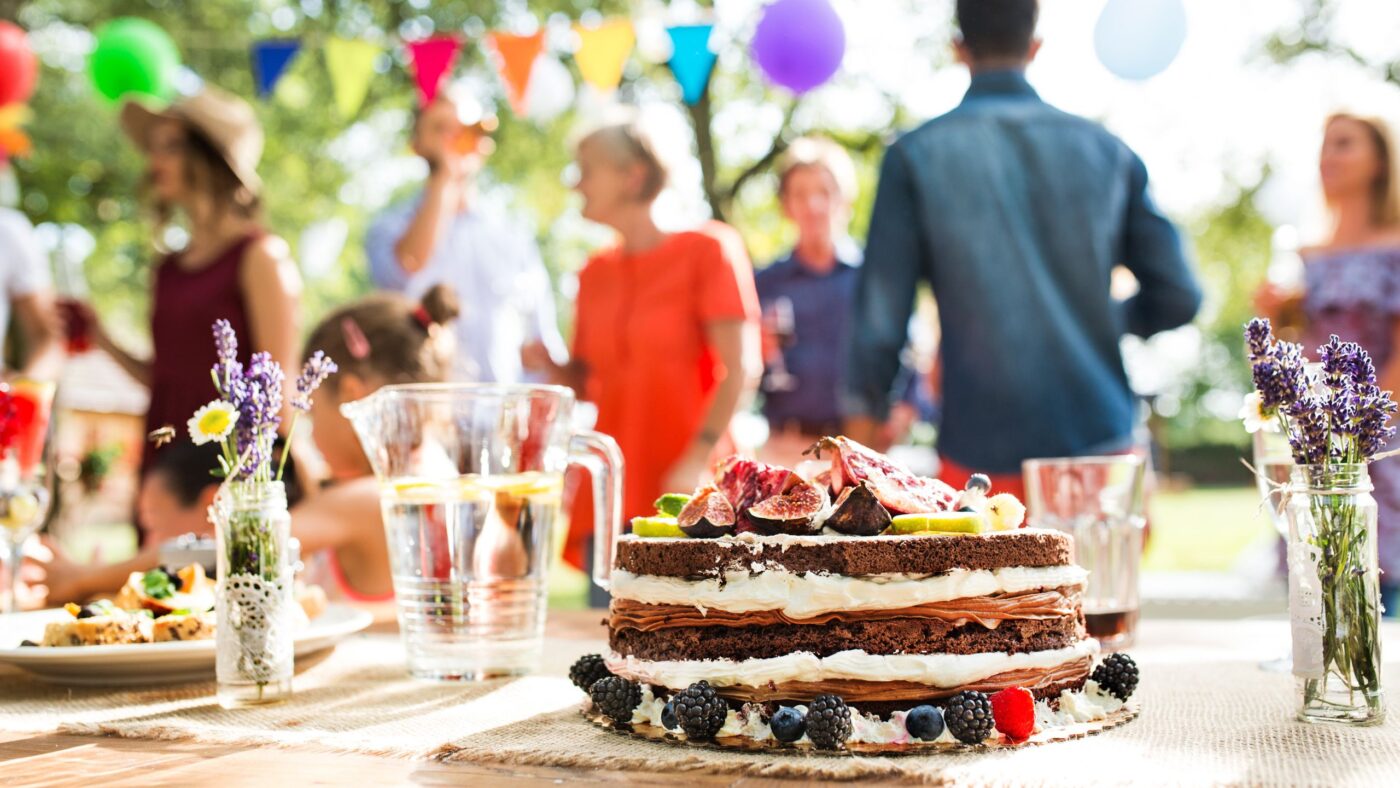Eine Veranstaltung in der Natur. Man sieht einen Kuchen im Vordergrund und im Hintergrund tanzende Leute