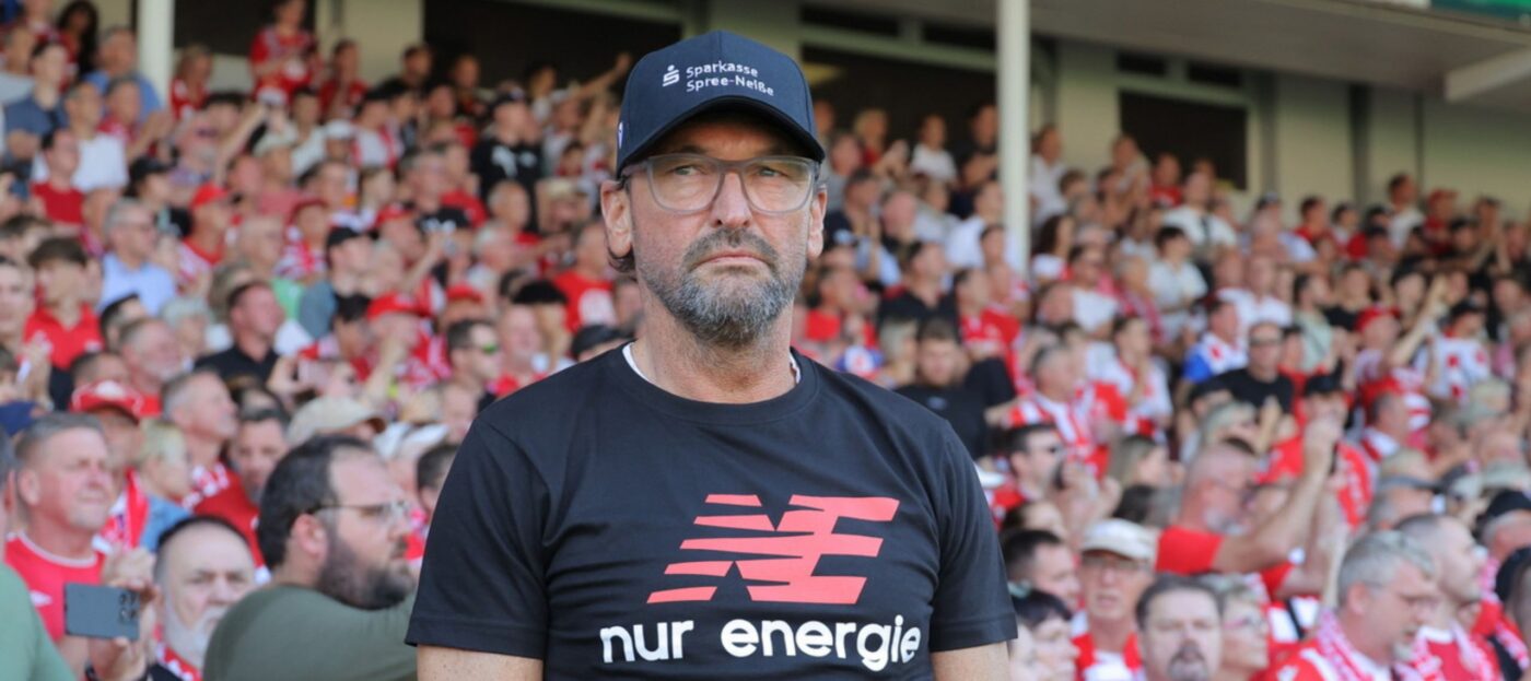 Claus-Dieter Pele Wollitz blickt skeptisch im LEAG Energie Stadion und steht vor der Tribüne mit vielen Fans im Hintergrund