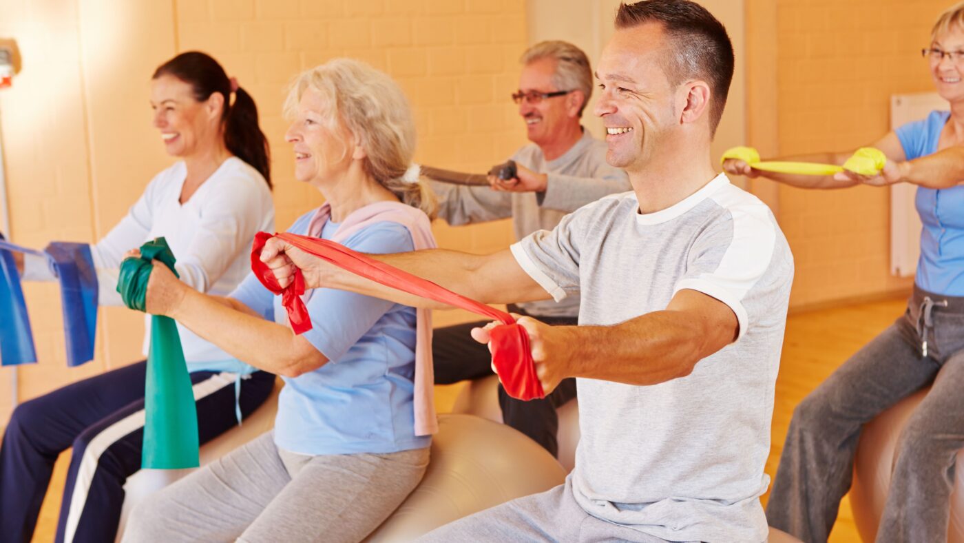 Eine Sportgruppe bestehend aus mehreren Männern und Frauen unterschiedlichstem Alter, die auf Gymnastikbällen sitzen und Sportübungen machen