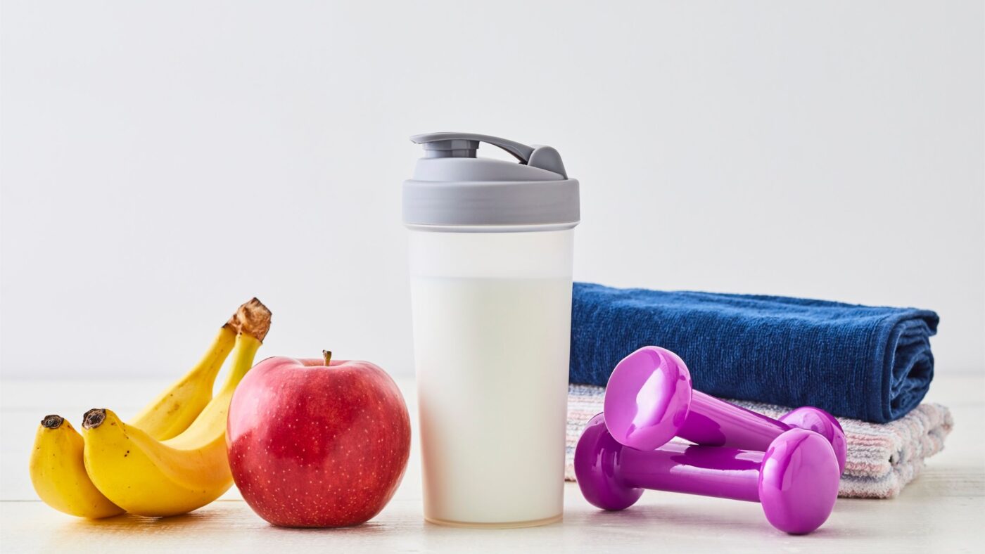 Das Bild als Darstellung zum gesunden Leben. Man sieht Bananen, Äpfel, ein Portein Shake und Sporthanteln