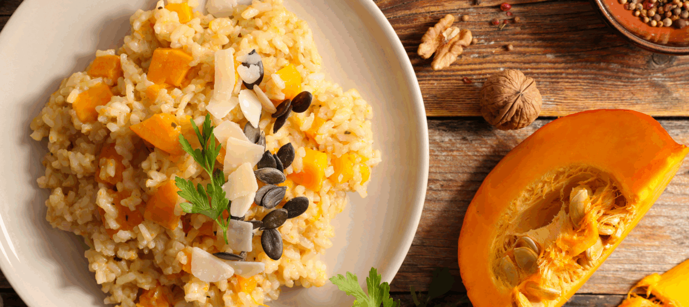 Ein auf einem weißen Teller angerichtetes Kürbisrisotto steht auf einem Holztisch im Herbst und daneben legt ein aufgeschnittener Kürbis