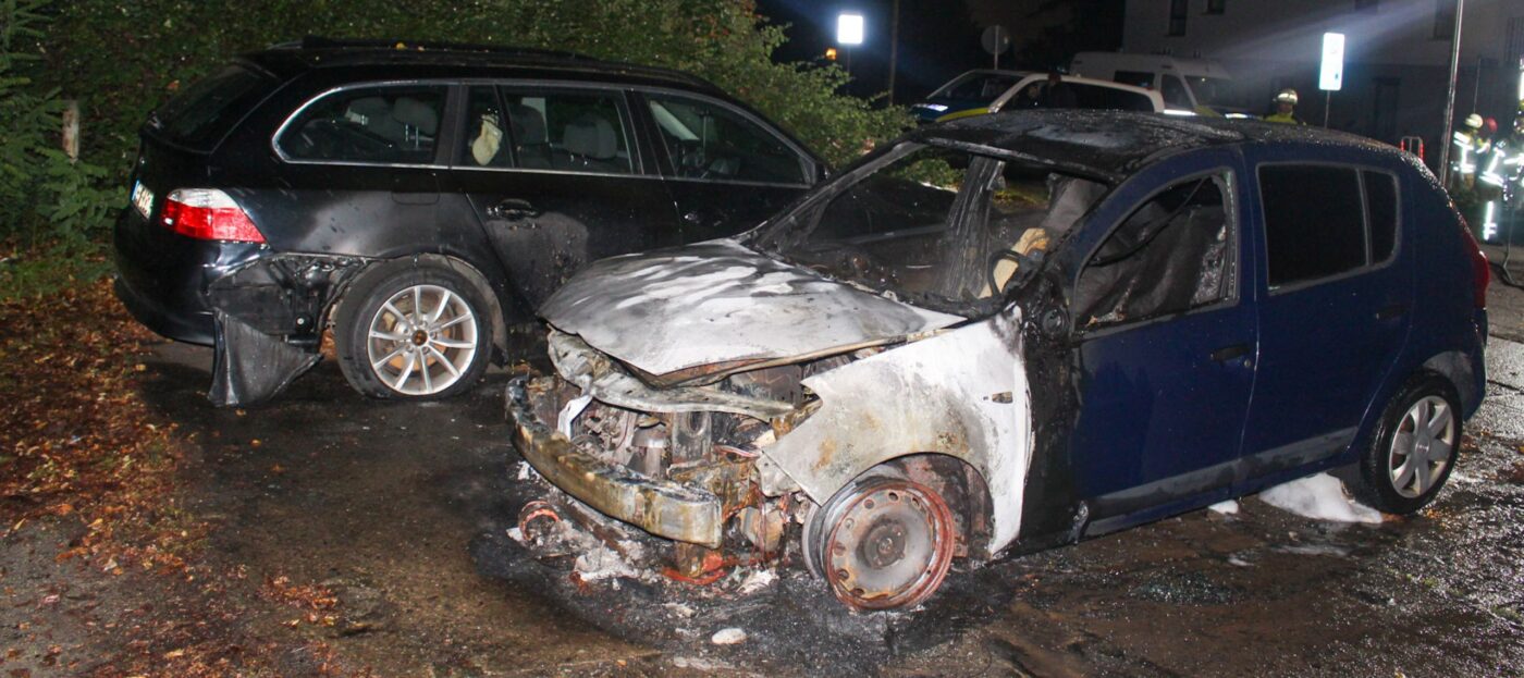 Ein vollkommen ausgebrannter PKW Dacia vor einem schwarzen BMW im Hintergrund, der ebenfalls Brandschäden am Heck zeigt