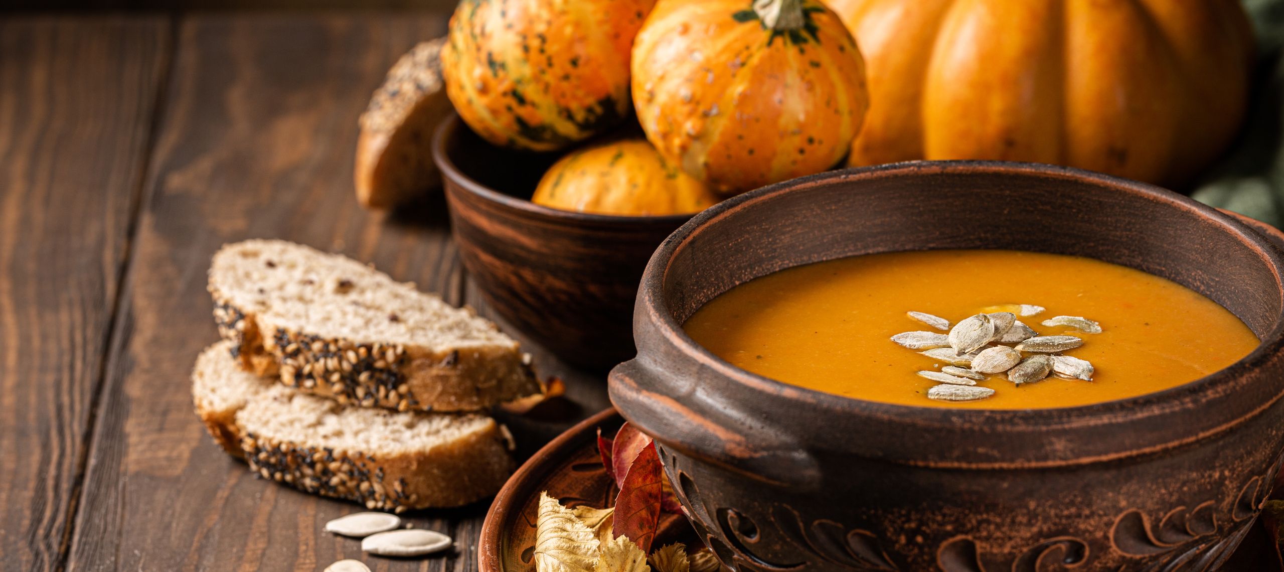 Kürbissuppe mit Kürbiskernen angerichtet in einer braunen Keramikschale mit Brot an der Seite und weiteren noch rohen Kürbissen im Hintergrund
