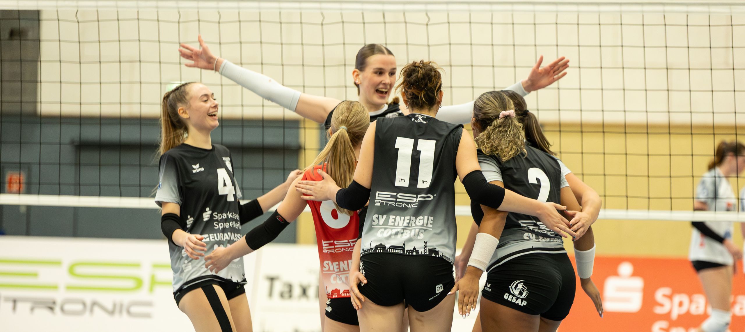 Die Mannschaft des SV Energie Cottbus jubelt in eigener Halle nach einem Punktgewinn gegen den Meister SCU Emlicheim mit weit ausgebreiteten Armen vor dem Volleyball Netz und will in Düsseldorf nachlegen