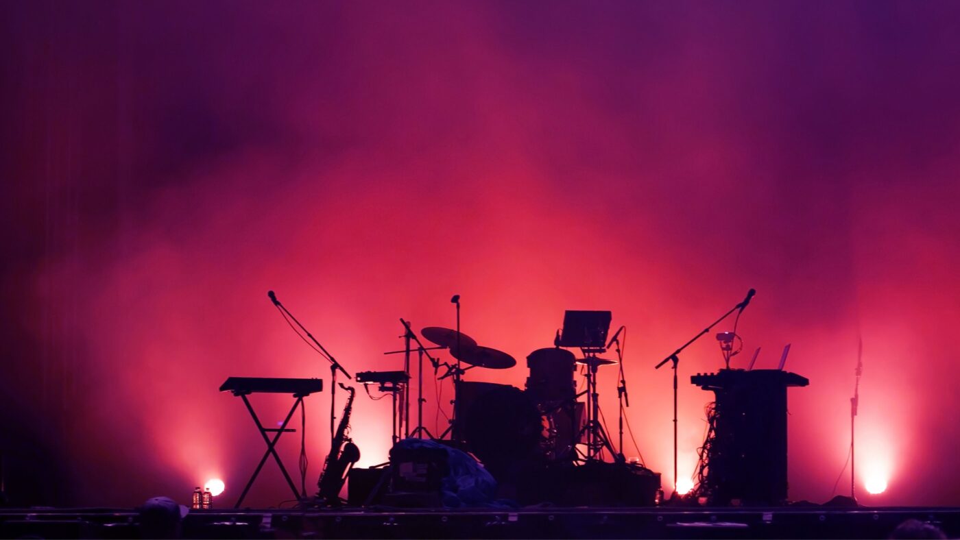 Bandinstrumente wie Gitarren und Schlagzeug auf einer Bühne, aber nur die Silhouette davon und dahinter rot-violettes Licht