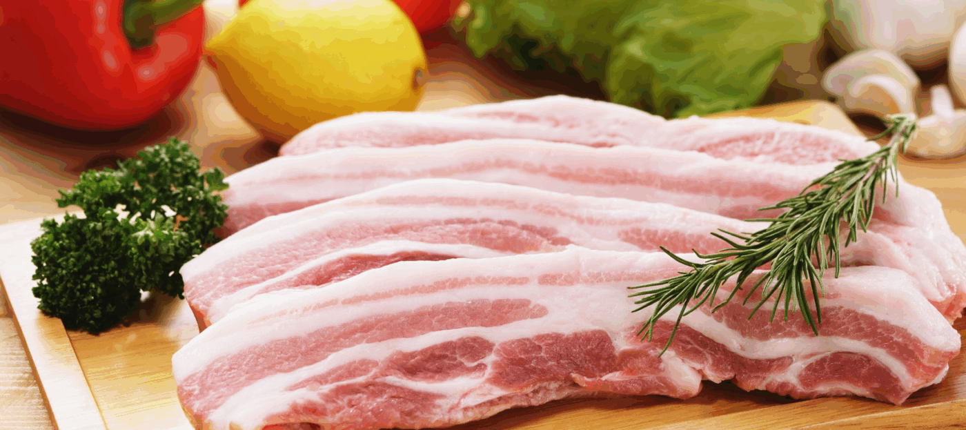 Ein Stück Schweinebauch liegt noch roh auf einem Küchenbrett im Hintergrund sind Paprika und ein Stück Herbst Kräuter liegt auf dem Fleisch