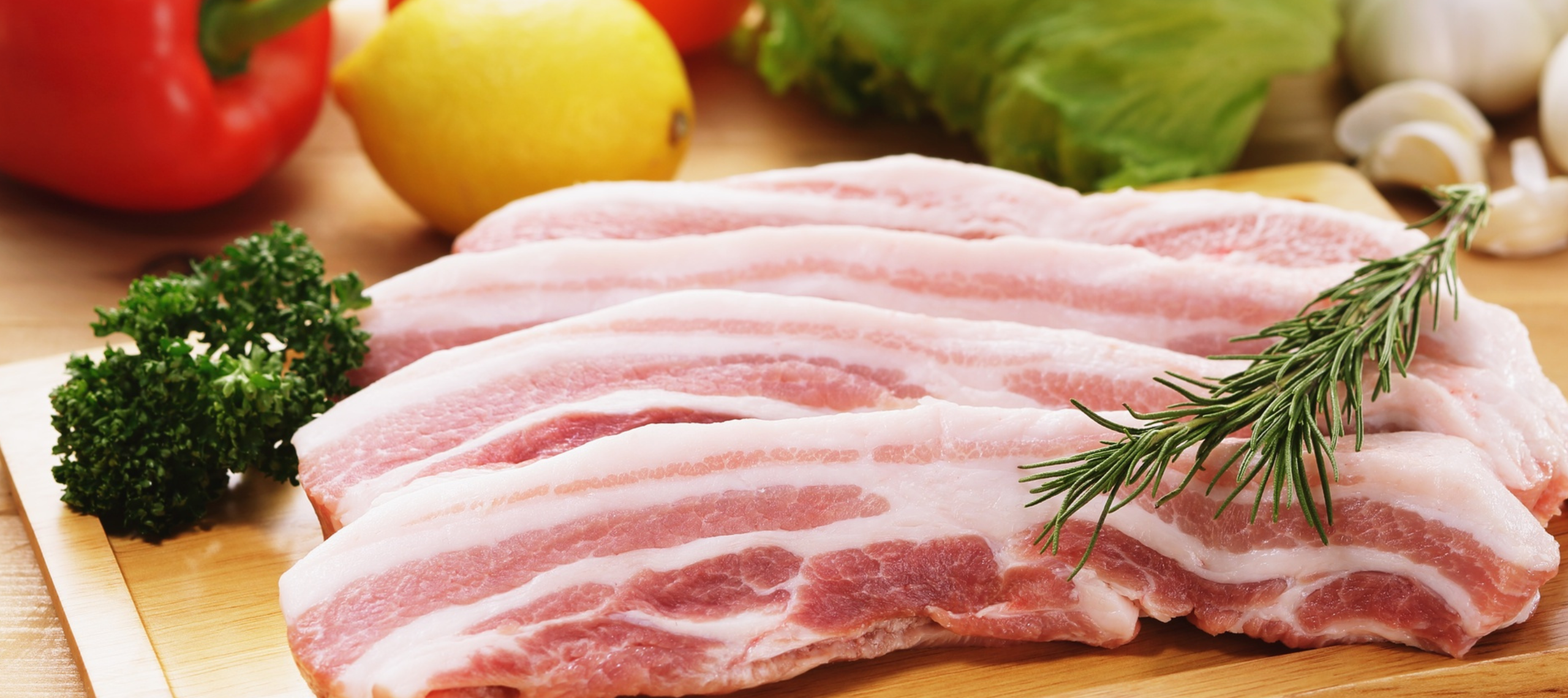 Ein Stück Schweinebauch liegt noch roh auf einem Küchenbrett im Hintergrund sind Paprika und ein Stück Herbst Kräuter liegt auf dem Fleisch