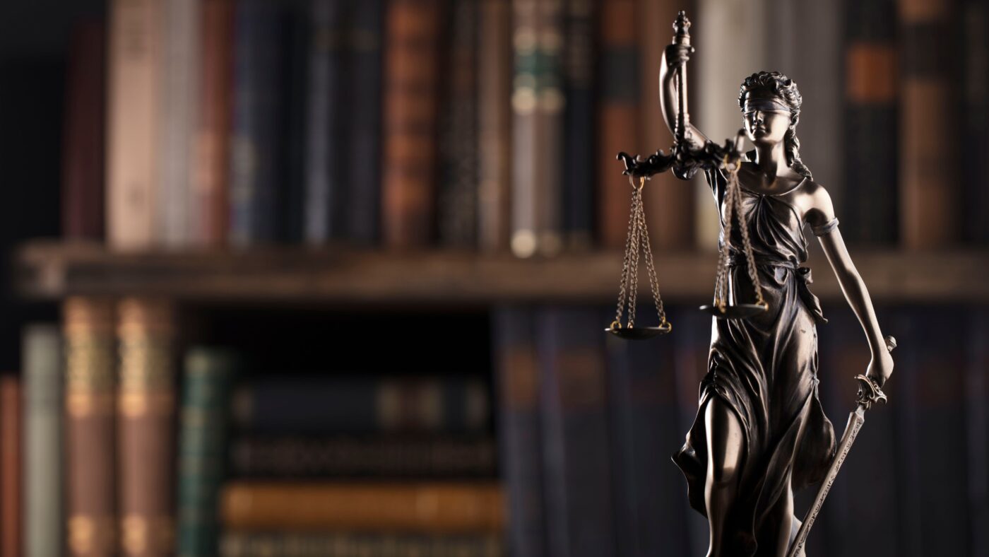 Eine Bronzefigur der Justizia vor einem unscharfen Bücherregal