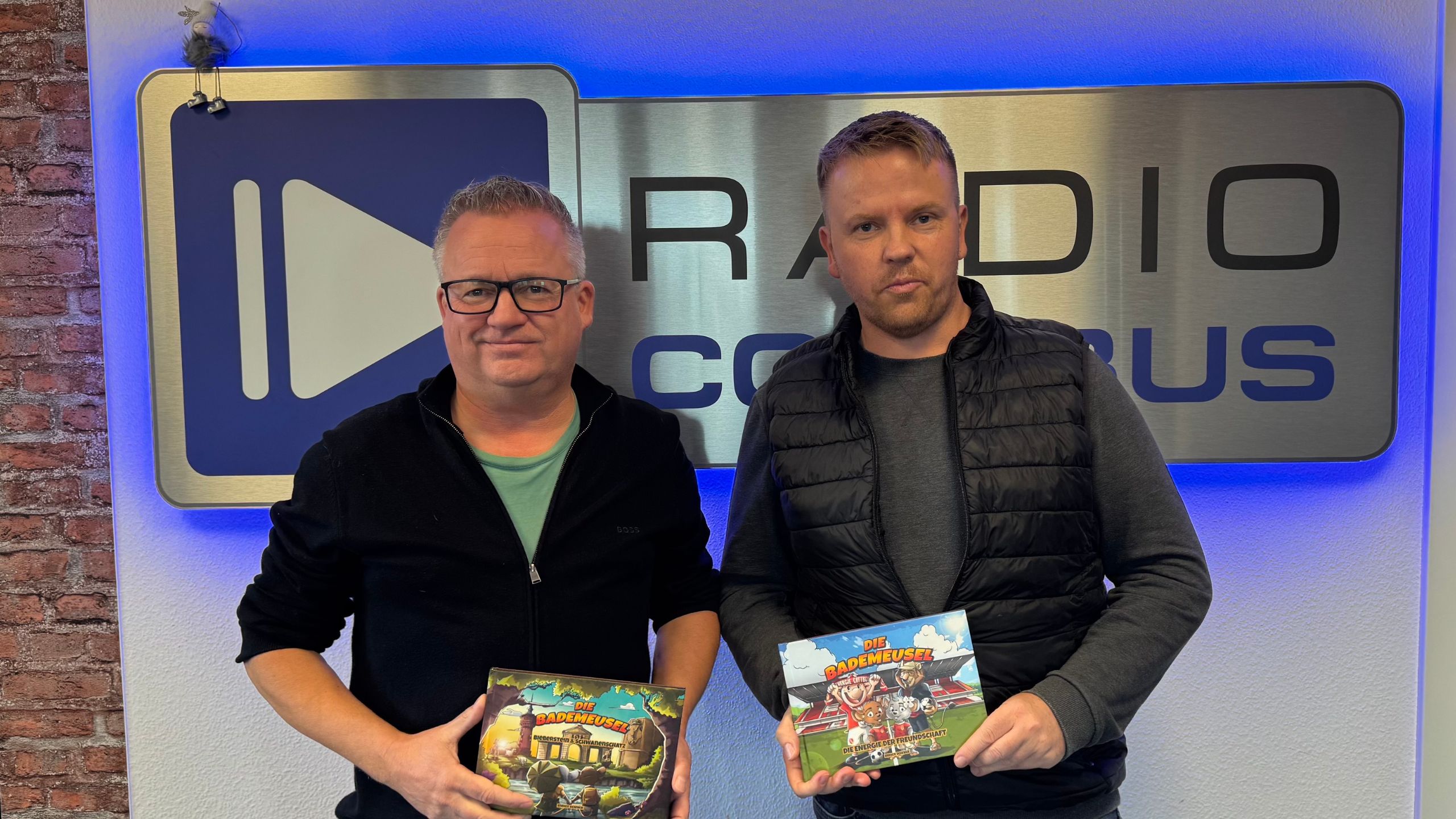 Enrico Schnick und Jörg Schröter halten das Buch "Die Bademuesel" vor dem Radio Cottbus Logo hoch.