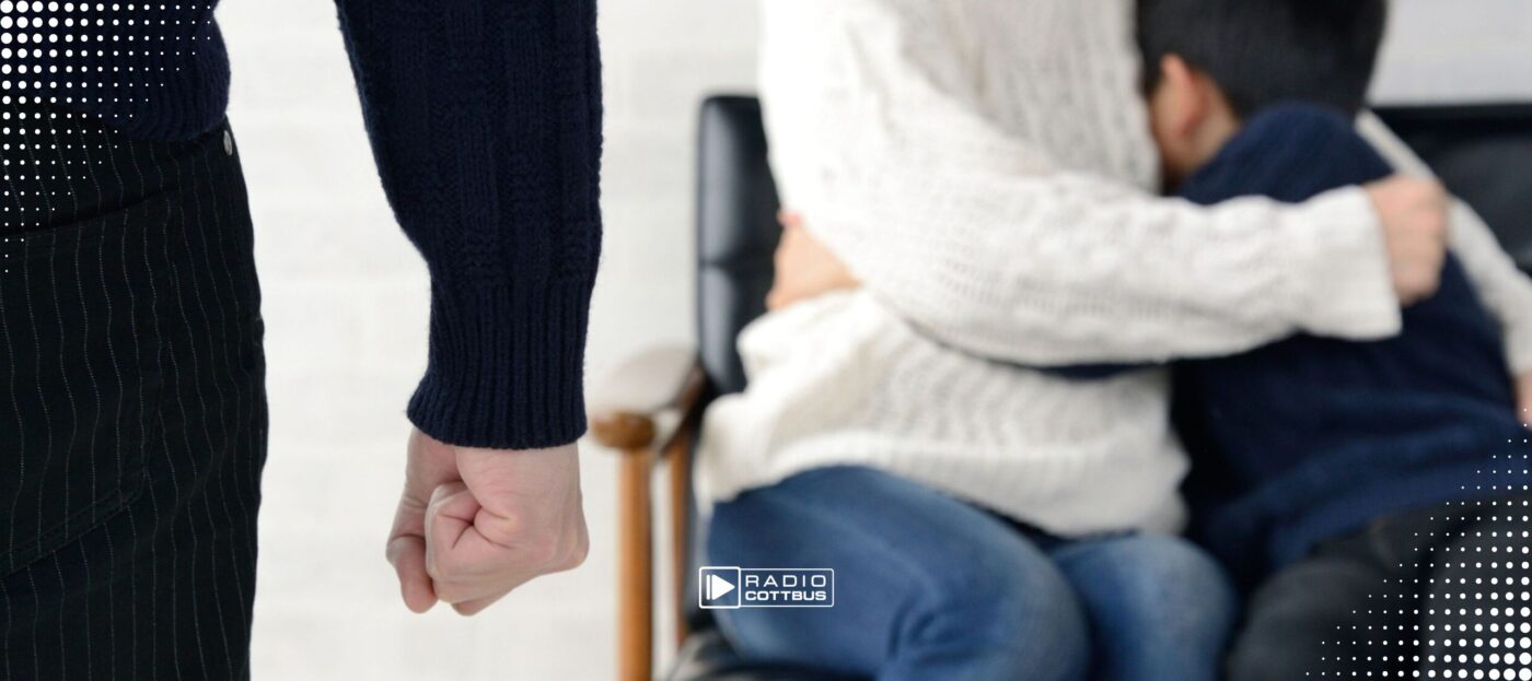 Links im Bild: ein Arm mit Faust, weiter rechts im Bild: eine Frau und ein Junge sitzen auf einem Stuhl und umarmen sich ängstlich, man sieht keine Köpfe oder Gesichter
