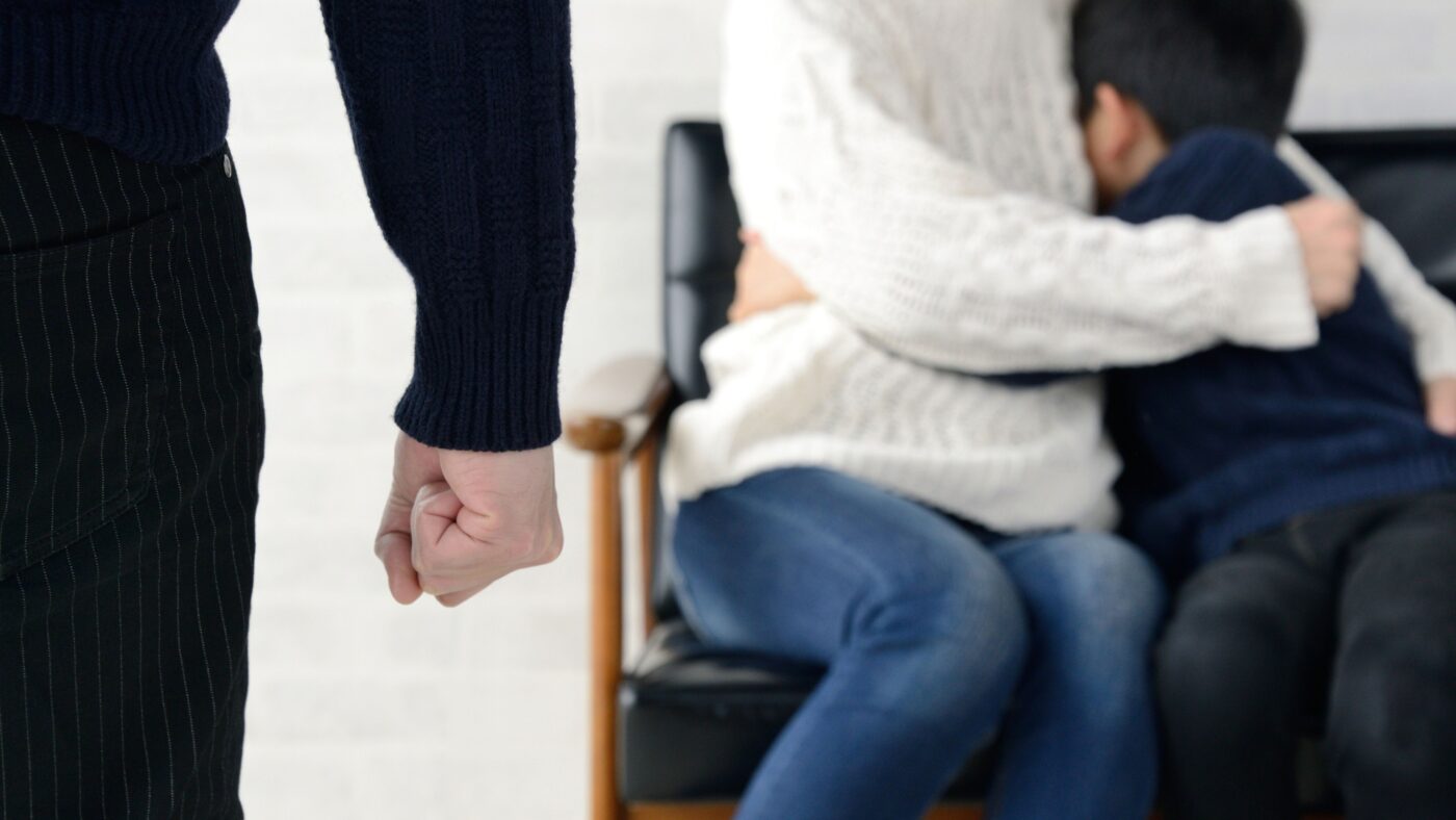 Links im Bild: ein Arm mit Faust, weiter rechts im Bild: eine Frau und ein Junge sitzen auf einem Stuhl und umarmen sich ängstlich, man sieht keine Köpfe oder Gesichter