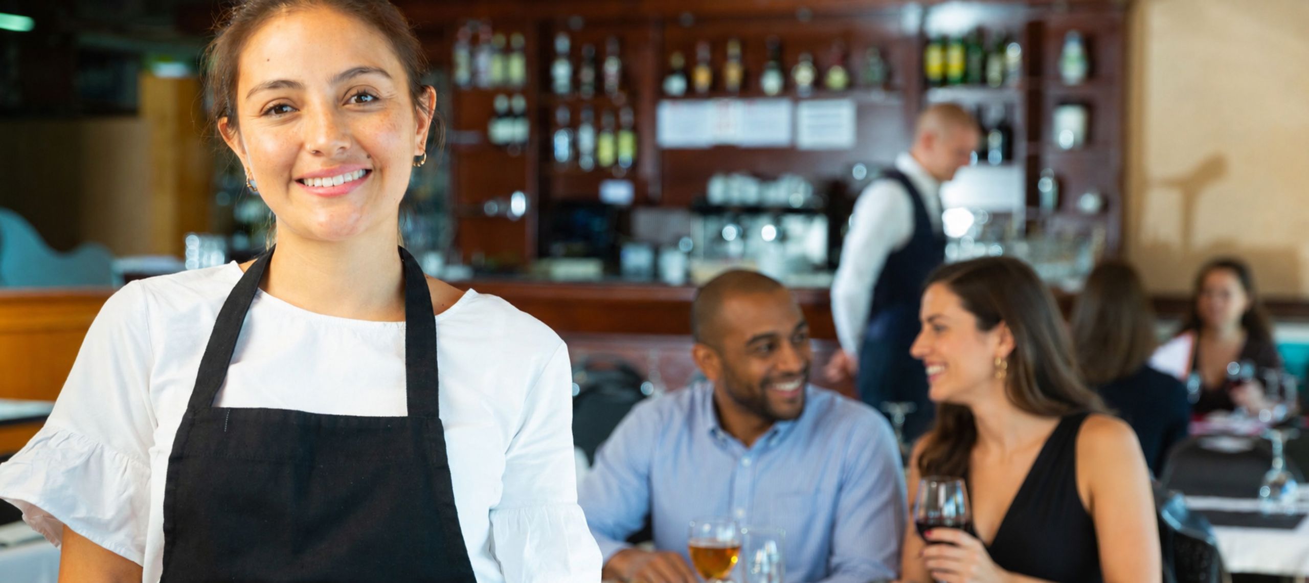 Eine Kellnerin lächelt in die Kamera in einem italienischen Restaurant in dem ein Pärchen im Hintergrund mit einem Bier und einem Glas Wein in der Hand sitzt und lächelt